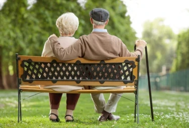 foto van achter gefotografeerd van een ouder stel dat samen op een bankje zitten in een park waarbij de man zijn arm op de schouder van de vrouw legt (relatie makend met duur van het leven)