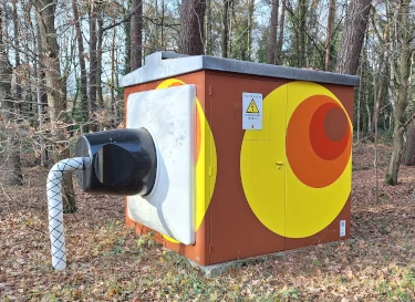 foto van een transformatorhuisje met aan de zijkant een enorm grote stekker, het transformatorhuisje is echt, de stekker is een aangebracht kunstwerk