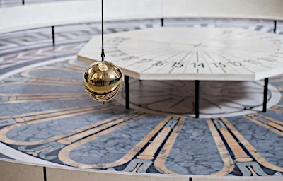slinger aan een lang touw die heel langzaam heen en weer beweegt over een tableau waar de uren van de dag staan en door de draaiing van de aarde de tijd weergeeft